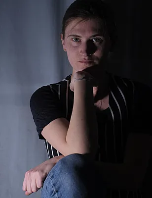 junge Frau, dunkelblond, grünäuig, in jeans, t shirt und gestreifter Weste, hockend, blickt mit auf Handrücken abgestütztem Kinn in die Kamera. im Hintergrund ist ein hellblauer Vorhang, der Bildausschnitt zeigt nicht die vollständige Person.